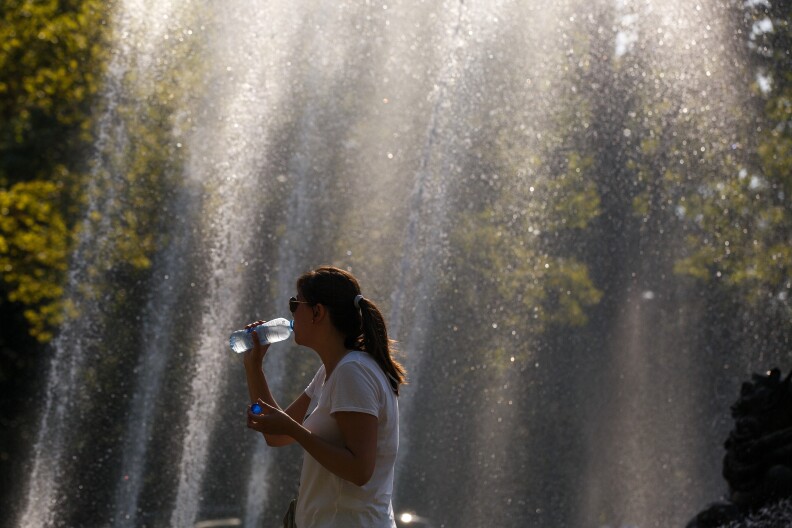 A woman with a brown ponytail and a white t-shirt sips from a plastic water bottle. She is taking is mist from a fountain. Green tree leaves are visible in the background.