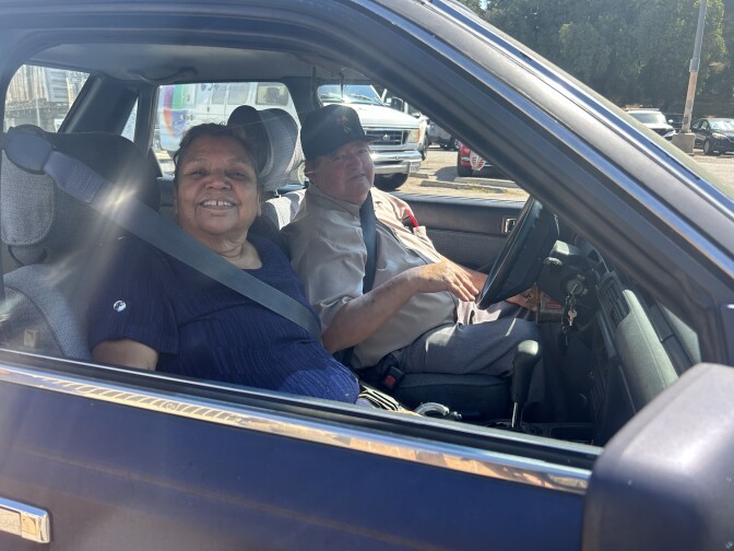 An older couple sits in a car smiling. The woman is on the left and wears a blue shirt and the man is on the right wearing a black baseball cap and khaki button down shirt. 