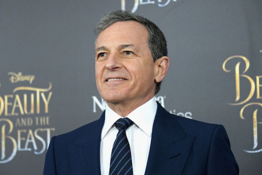 A light skinned man with greying hair stands in a blue suit with shirt and tie before a backdrop which reads "Beauty and the Beast."