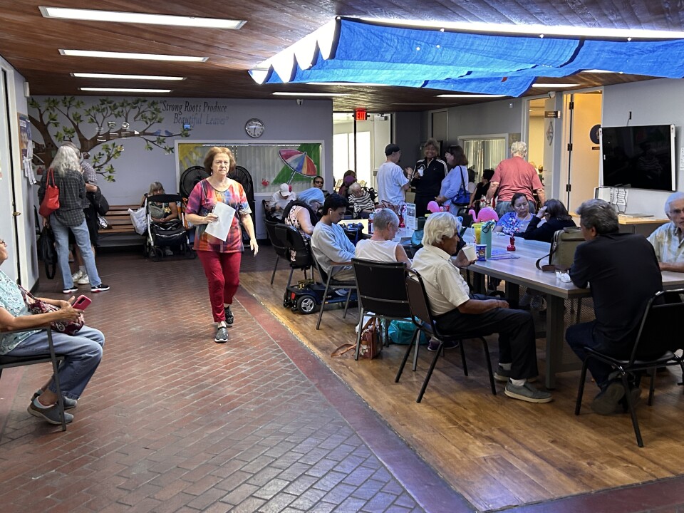The interior of a senior center, with about a dozen older adults gathered around a rectangular table in the center of the room.