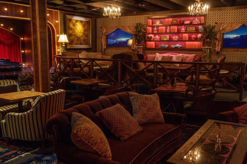 A lounge with plush, colorful furniture, carpeting and a small stage with a piano in front of a red curtain.