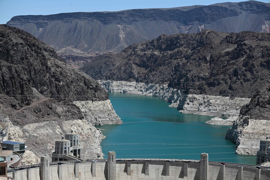 The rocky banks of a river show a white band toward the bottom, indicating a previously higher water level. The Hoover Dam is shown in the foreground.