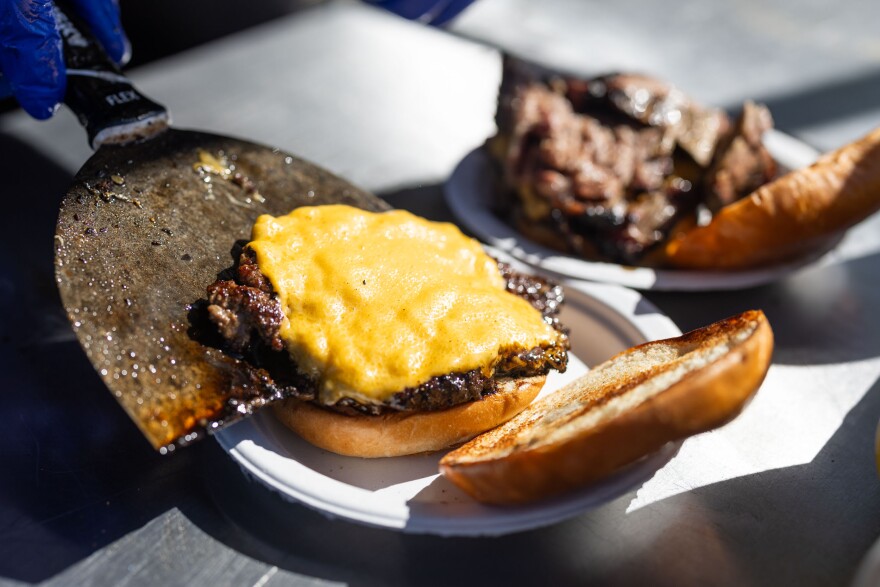 A close up of a burger patty with melted American cheese on top being placed on a bun that sits atop a aper plate. A second burger is in the background.