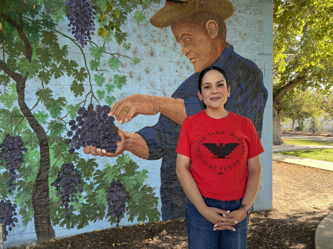 A woman with medium-light skin tone and dark hair that's pulled back into a ponytail is clad in a red t-shirt with a black emblem of an eagle. The t-shirt reads: "United Farm Workers" and "¡Sí se puede! in Spanish. She smiles gently while standing before a mural of a farm worker harvesting grapes. 