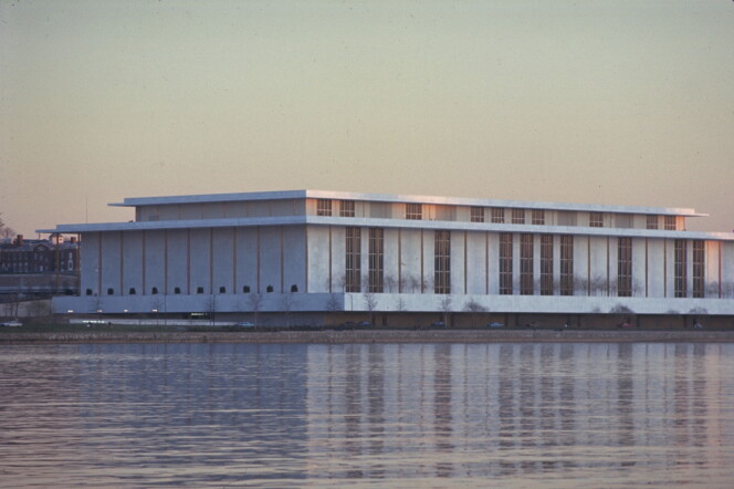 A large, low white building sits on the bank of a river. It is pictured from across the river.