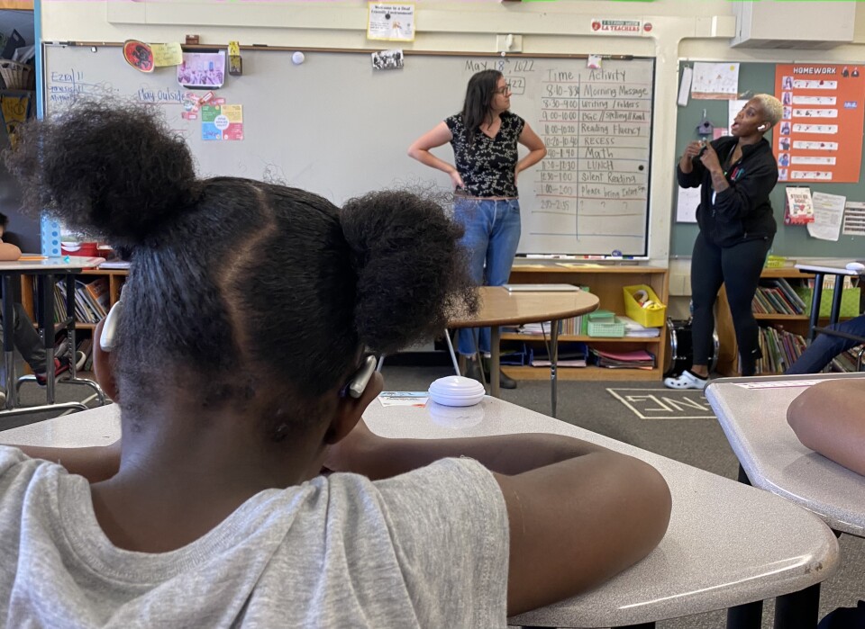 In the foreground of the image, an elementary-aged student seated at a desk in a school classroom faces away from the camera. Her hair is pulled into two pigtails. Assistive hearing devices are clearly visible hooked over the top of her ears. In the background of the image — the front of the classroom — a teacher and an aide speak to each other in American Sign Language (ASL).
