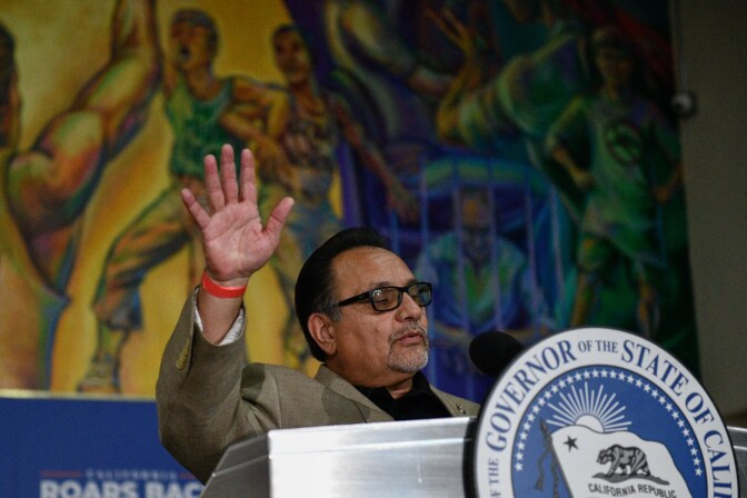 A man wearing glasses with his right hand raised stands at a lectern with an emblem of the governor of California.