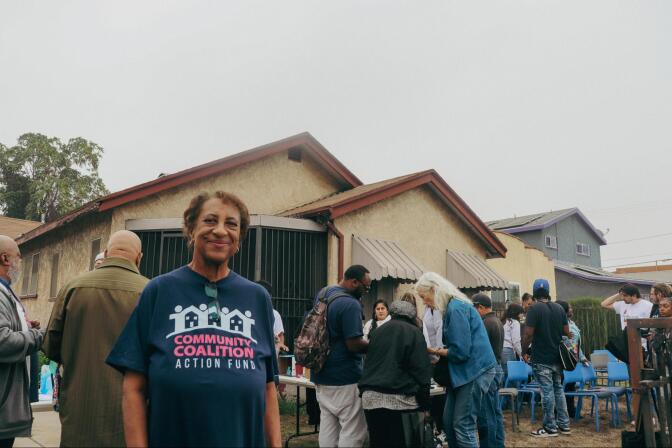 A smiling woman wearing a blue shirt that reads "community coalition action fund" stands in front of a beige house. Behind her, a group of people chat around a table and rows of blue plastic chairs.