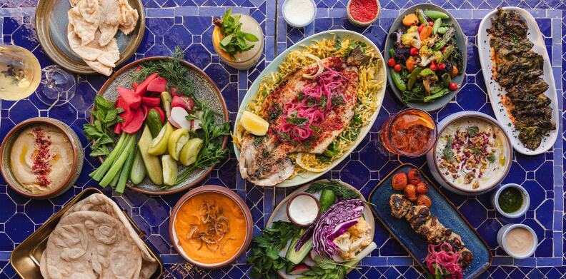 A blue, tiled table is set with a variety of prepared dishes, including skewers, salads, fish and hummus.
