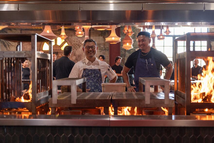 Two chefs, one in a white shirt and one in black, stand behind smile as they stand behind a professional stove with big flames going.
