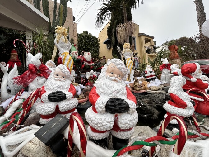 A series of Santa figurines, with their black-gloved hands folded in front of them, are displayed in an outdoor space. 