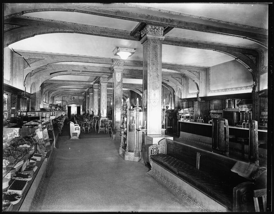 A sweets showcase can be seen at left, while long benches for waiting can be seen across the showcase at right. A bar with a few stools in front of it can be seen at right behind the bench. Tables and chairs can be seen in the background, while wooden beams are visible on the ceiling. Decorative pillars are visible down the center of the room.