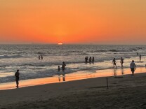 The sun sets over the ocean and people stand in the water.