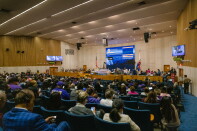 A large auditorium where a large crowd sits in blue chairs facing a dais with a large screen above. 