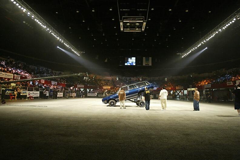 A blue station wagon is raised up on its front tires in a stadium as lights shine on the car.