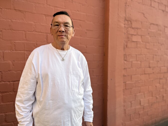 A bespectacled Asian man in his late 60s wears a white shirt and gold chain around his neck.