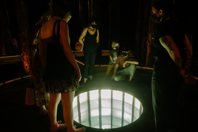 A group of people wearing masks stands around a circular, illuminated hole in the floor. It appears to continue down into infinity, due to an optical illusion.