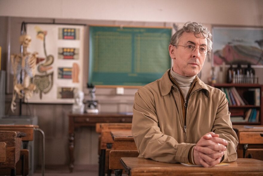 Alan Cummings sits in a desk in a classroom on the right side of the frame. He wears a tan jacket zipped halfway up, with another brownish jacket underneath zipped slightly more up, with a turtleneck underneath it all. His hands are clasped together on the desk, and he stares out of his glass out of the screen. In the background: two desks behind Alan, with some more flanking him on either side; a table with a bust and microscope, a bookcase, and a skeleton with a poster of the human GI tract behind it.