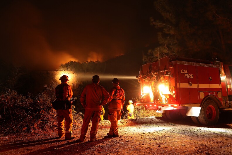 Firefighters watch a fire in the distance with a night sky full of smoke.