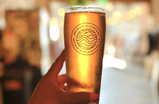A golden glass of beer has light coming through it. It's being held by a hand. On the glass it says Brewjeria Company, brewed in Pico Rivera