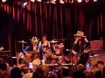 Dwight Yoakam plays to a crowd at The Roxy. He plays a acoustic guitar and wears a blue jacket and white cowboy hat. 
