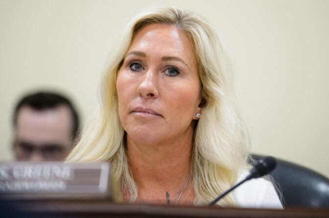 A woman with light-tone skin and blond hair sits in front of a mic and name card that's blurred.