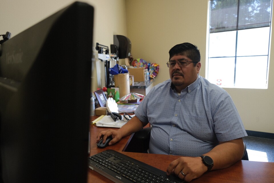 A man with medium-tone skin is in a short-sleeved button up shirt and sits at a desk working on a computer.