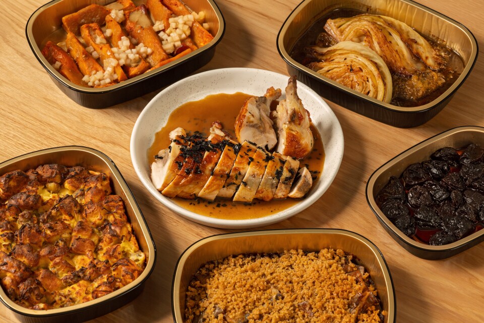 Top-down photo of a sliced Chicken Ballotine — heritage chicken breast stuffed with chicken, Chinese sausage, shiitakes, ginger, chestnut, and black truffle—covered in gravy on a white plate. Surrounding it on a wood table are trays of roasted cabbage, sweet potatoes, a crumb-topped casserole, baked stuffing and dark glazed prunes.