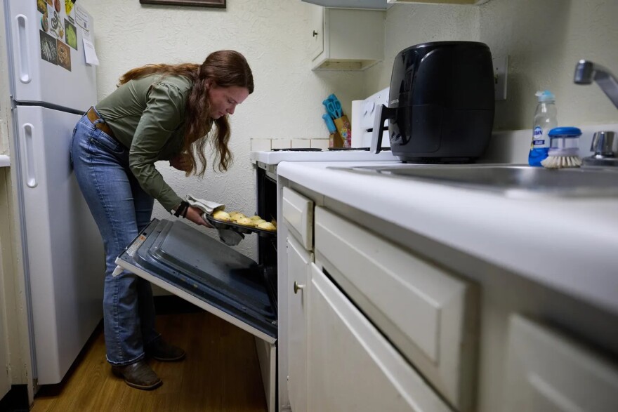A woman wearing jeans and a green shirt pulls out a muffin pan from an oven