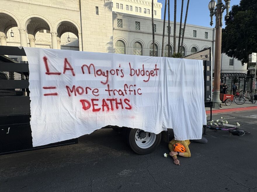 A man with an orange helmet lies in the road behind a truck with a white rose in his right hand. The truck is draped with a white sheet bearing red text: "LA Mayor's budget = more traffic DEATHS."