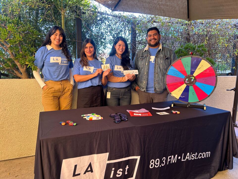 Four people wearing a light blue t-shirt with a logo that reads "LAist" pose for a photo. Two hold a sticker and bookmark as the group stands in front of a table with a black tablecloth that reads "LAist. 89.3 FM. LAist.com" with a spinning prize wheel and other items on top of the table.