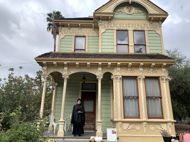 A green Victorian home at Heritage Square Museum. A woman in mourning clothes stands on the front porch.