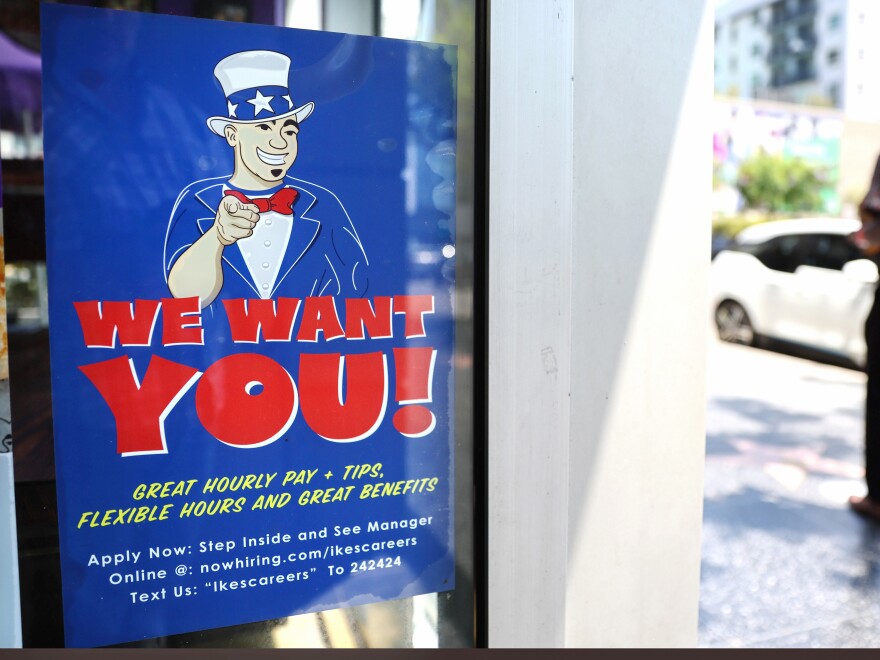 A large sign taped in a store window reads "We Want YOU! Great hourly pay + tips, flexible hours and great benefits."