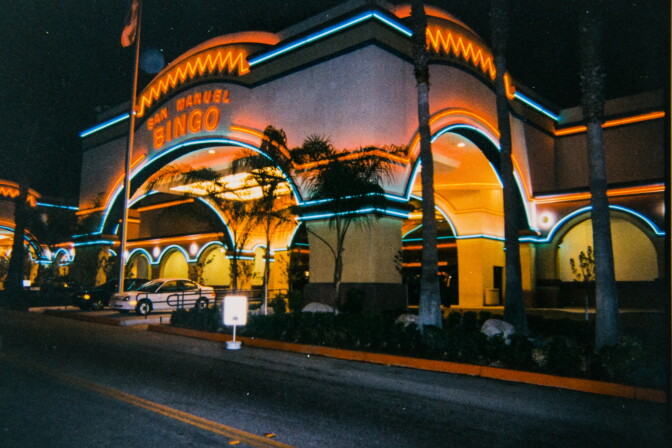 A wide view of San Manuel Bingo from the front entrance. Some cars are in front. The building facade has curved entryways with red and blue neon accent lighting.