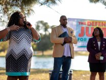 LA County Superior Court judge candidates La Shae Henderson, George Turner and Ericka Wiley speak at a Defenders of Justice campaign event.  They stand on the grass in front of a body of water. A 'Defenders of Justice' campaign banner can be seen behind them. 
