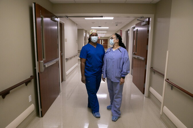 Two nurses, one in deep blue scrubs and the other wearing light blue, stand in a hospital corridor with masks on.
