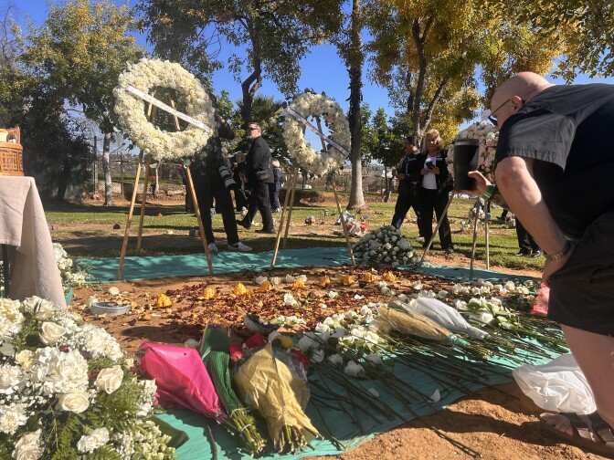 A man wearing all black t-shirt and shorts is leaning over a large communal grave buried in the dirt. He has a black coffee cup in his hand, outstretched towards the grave as if in a toast. The grave is covered in white roses and flower bouquets. 