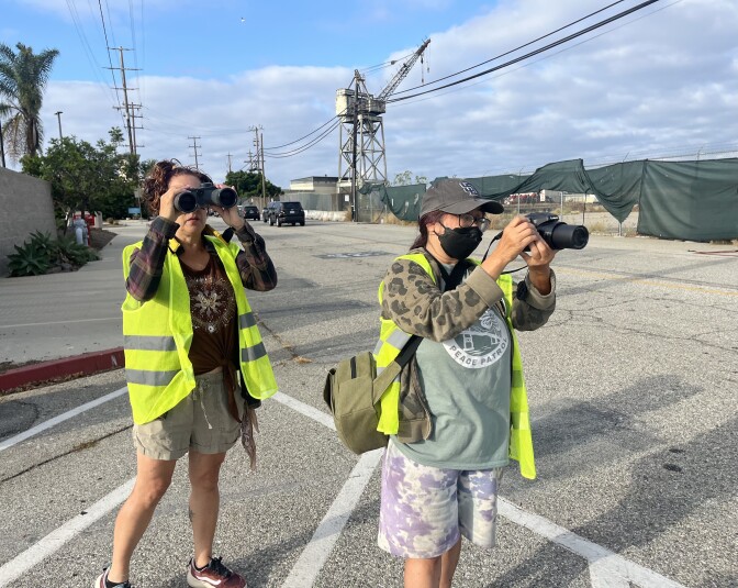 Two women in yellow safety vests stand on a street, looking through binoculars and a camera.