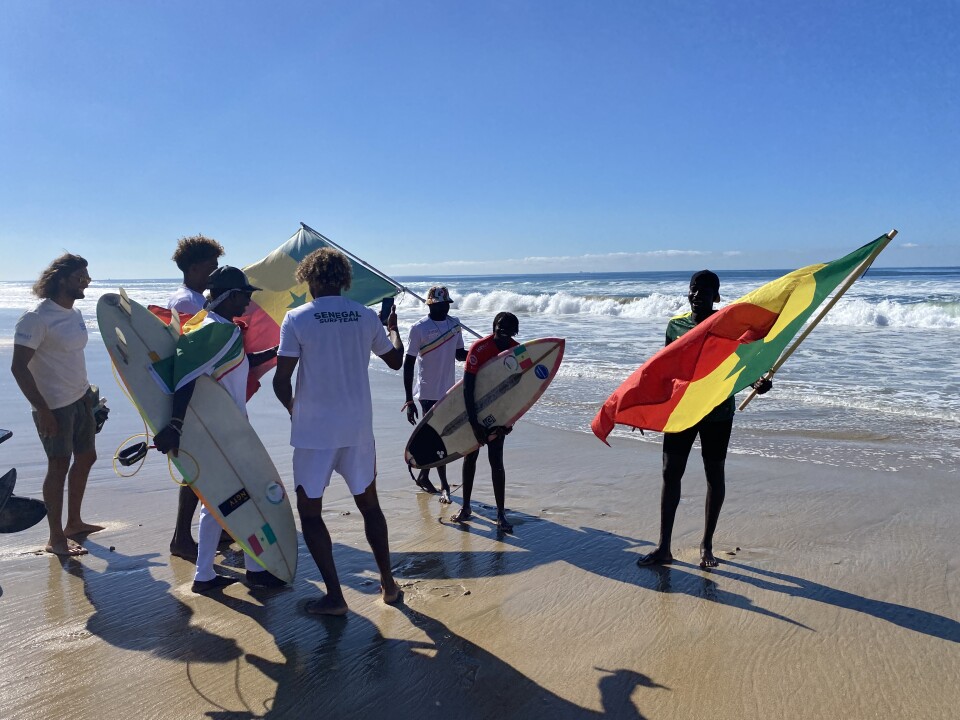 Six men in white t-shirts, two of them waving red, yellow and green Senegalese flags, greet a Black woman with short hair, wearing a red jersey and holding a surfboard on the beach. 