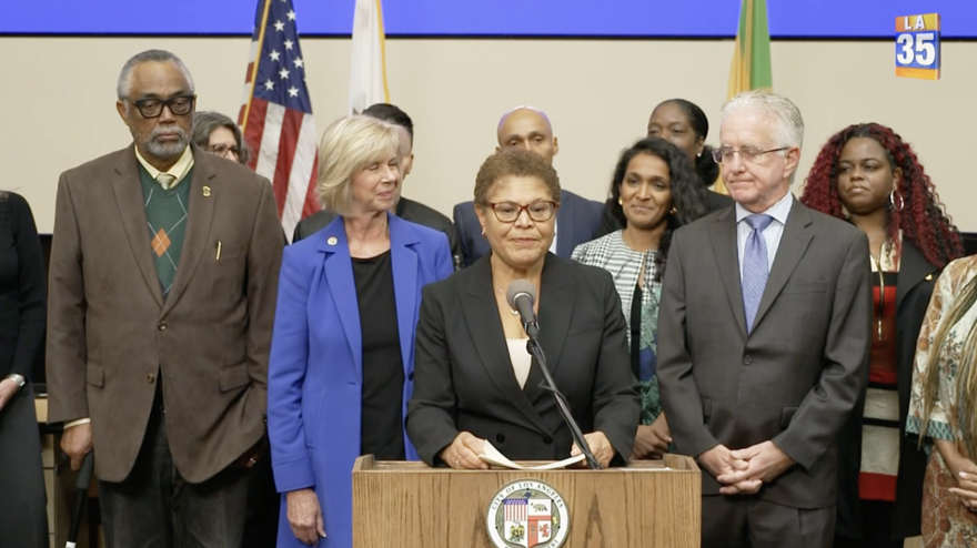 Mayor Karen Bass stands at a podium, backed by a diverse group of city officials. She is wearing a black suit jacket and glasses.