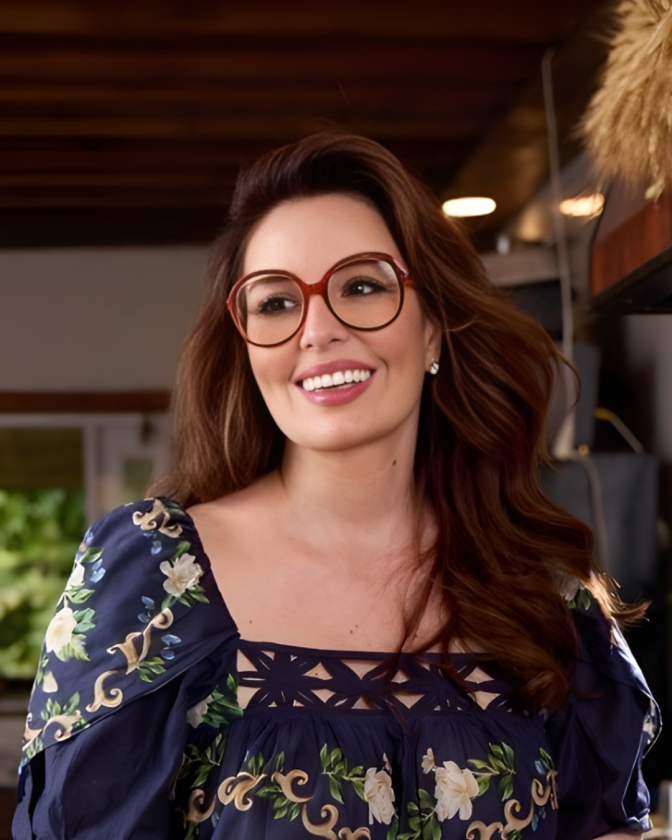 A woman with a light skin tone smiles in a kitchen setting, wearing large tortoiseshell glasses and a floral navy dress.