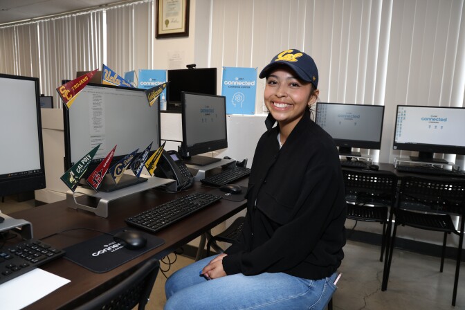 A young woman with medium toned skin is sitting in front of a desktop computer that's decorated with various college flags. She's wearing a black jacket and a baseball cap from UC Berkeley.  