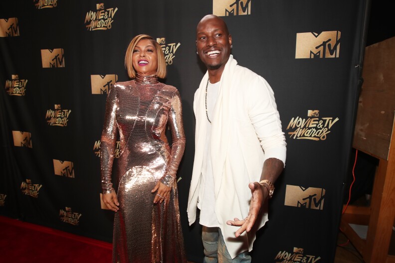 A Black woman wearing a shiny silver dress and a Black man dressed in white are on the red carpet looking off-camera and smiling.