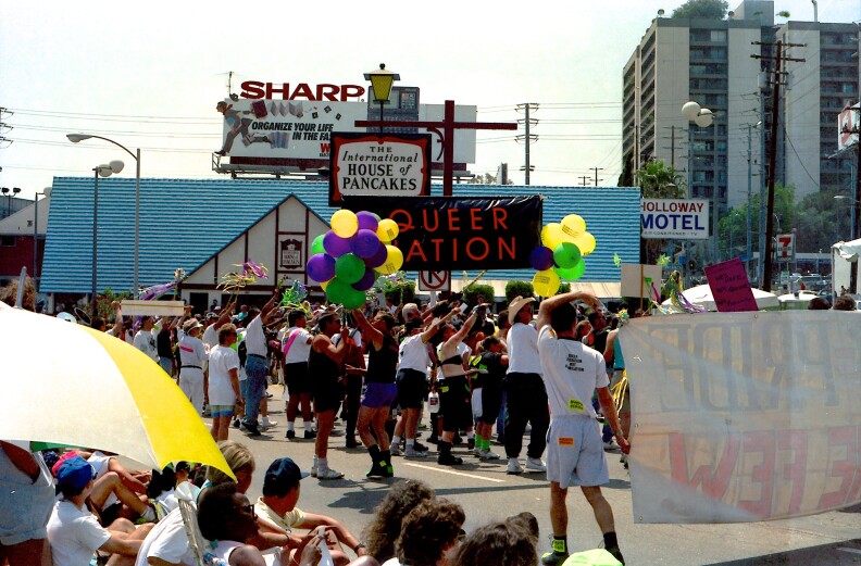 A view of a crowd on the street during Pride. A group is holding a black banner in the distance with red letters that say queer nation.