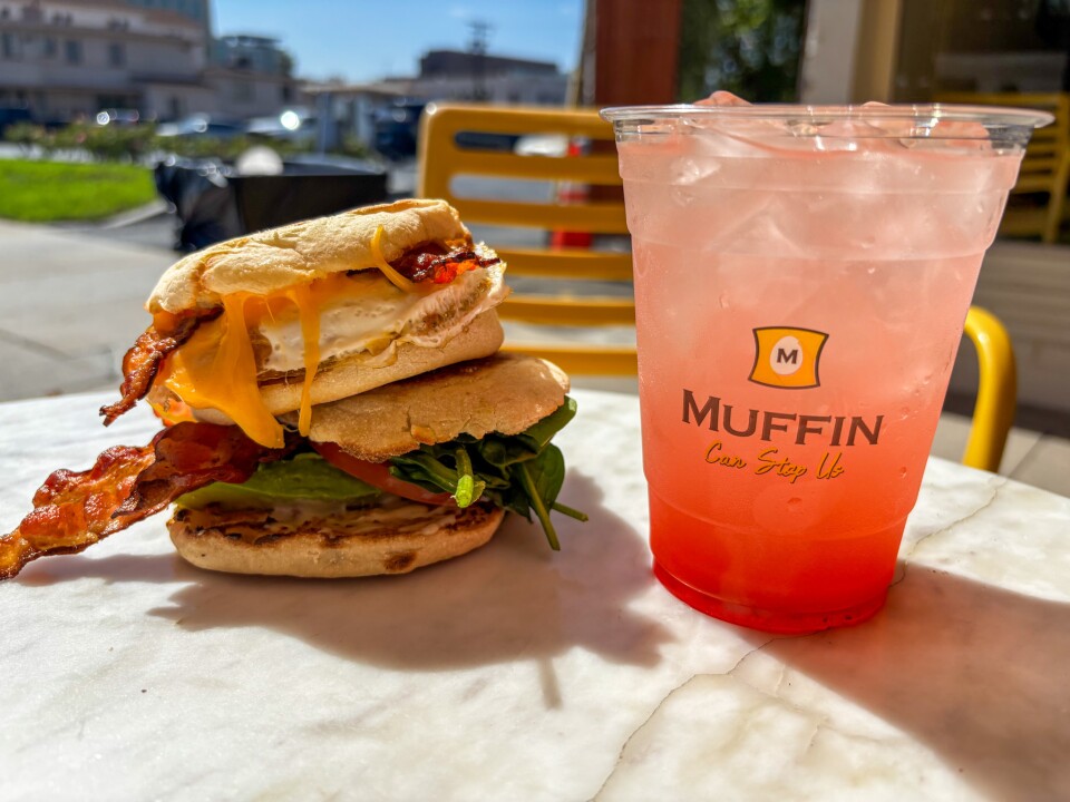 Two breakfast sandwiches sit piled on top of each other, both made with English muffins. One is with bacon and melted cheddar cheese. The other has bacon, lettuce, tomato, and egg. Both are paired with a cold pink lemonade in a clear cup labeled "Muffin Can Stop Us."