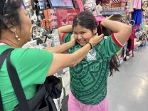 A young, female presenting person holds her arms up and wears a green sports jersey. Her mother holds her cheeks in a caring way.
