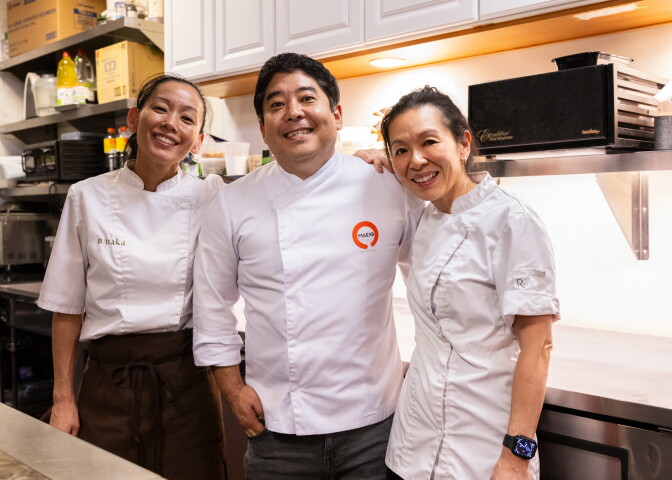 Three smiling chefs in white jackets stand shoulder to shoulder in a kitchen with their arms around one another. Carole Iida-Nakayama, a woman, on the left, has medium-light skin and dark hair pulled back, and wears a jacket stitched “n/naka” and a brown apron. Micha Tsumura, a man, in the center, has medium-light skin and short black hair, and wears a jacket with an orange Maido logo. Niki Nakayama, a woman, on the right, has medium-light skin and dark hair pulled back, and wears a smartwatch.