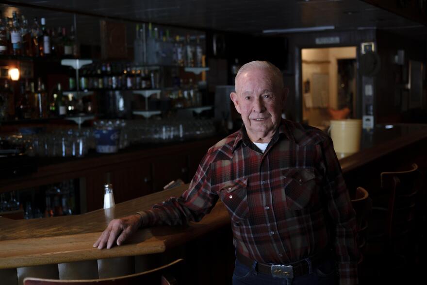 A wide shot of a man in a plaid shirt and blue jeans standing inside an empty bar, next to a wooden bar