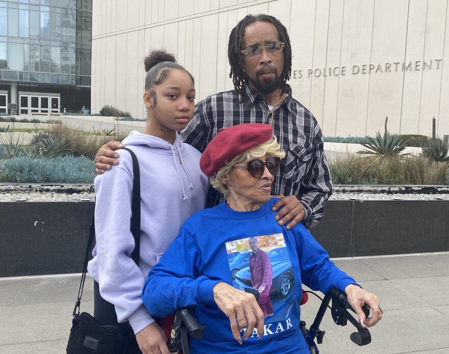 Takar Smith's family comes together for a photo outside LAPD headquarters on Friday, January 13. Smith's niece, Rashi wears a purple hoodie. His brother, Raischard, wears glasses and a plaid shirt. Raischard has his hand on their mother, Alicia Smith, who wears a blue shirt that has an image of Takar on it.