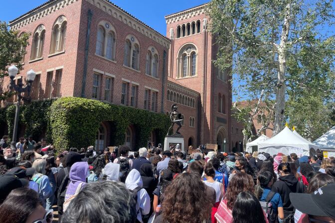 A large group of people with their backs mostly towards the camera stand in front of USC's Tommy Trojan statue. A red brick building with beige windows is visible in the background. 